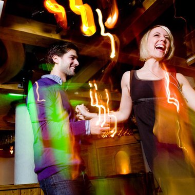 A couple dances happily in a lively club. Colorful lights and a festive atmosphere surround them. | © Aparthotel Kleinwalsertal Haller GmbH & Co KG