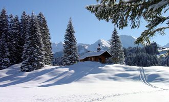 © Bruder Klaus Kapelle | Rolf Koeberle A snowy landscape with tall fir trees and a small wooden house. In the background, the snow-covered mountains and a clear blue sky can be seen. | © Bruder Klaus Kapelle | Rolf Koeberle