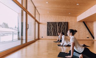 © Naturhotel Chesa Valisa A bright yoga room with wooden paneling and large windows. Three people are practicing yoga and looking outside. | © Naturhotel Chesa Valisa