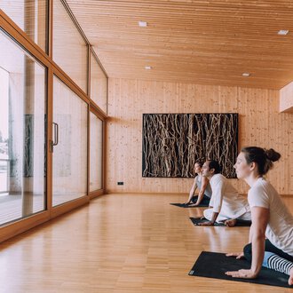 © Naturhotel Chesa Valisa A bright yoga room with wooden paneling and large windows. Three people are practicing yoga and looking outside. | © Naturhotel Chesa Valisa