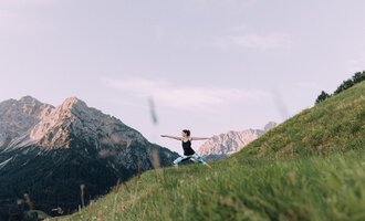 © Naturhotel Chesa Valisa A person is practicing yoga in nature on a green meadow. Impressive mountains are visible in the background. | © Naturhotel Chesa Valisa