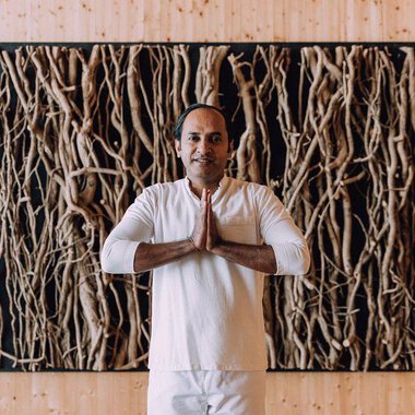 © Das Naturhotel Chesa Valisa A man stands with his hands folded in front of a wall made of natural materials. He is wearing a simple, light outfit and exudes calmness. | © Das Naturhotel Chesa Valisa