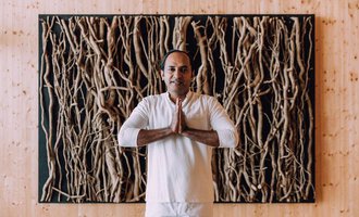 © Das Naturhotel Chesa Valisa A man stands with his hands folded in front of a wall made of natural materials. He is wearing a simple, light outfit and exudes calmness. | © Das Naturhotel Chesa Valisa