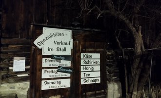 Ein Holzgebäude mit einem Verkaufsschild für Spezialitäten im Stall. Die Beschilderung weist auf Öffnungszeiten und angebotene Produkte wie Käse und Honig hin. | © Abler Spezialitätenverkauf im Stall | N. Lughammer