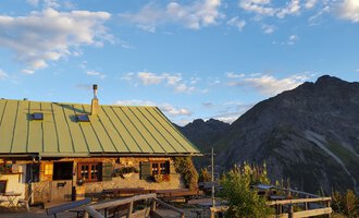 A cozy mountain hut with a green roof in the Alps. In the background, majestic mountains stretch out under a blue sky. | © Alpe Kuhgehren | Lisa Hiesinger