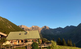 A picturesque alpine hut surrounded by mountains. The clear sky and the green meadow give the scene a peaceful atmosphere. | © Alpe Kuhgehren | Lisa Hiesinger