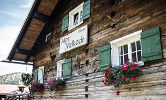 Ein traditionelles Holzhaus mit grünen Fensterläden. Blumenkästen mit bunten Blumen schmücken die Fenster. | © Kleinwalsertal Tourismus | Andre Tappe