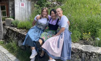 Three women are sitting on a stone wall in traditional attire. In the background, there is a green meadow and a wooden house. | © Alpe Widderstein | Tom Egger