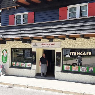 Ein traditionelles Restaurant mit einem einladenden Eingang und einer Terrasse. Die Fassade ist in hellen und dunklen Farben gestaltet, mit einer kleinen Gruppe von Menüs und Angeboten auf Plakaten. | © Alpengrill Riezlern | Veronika Senn