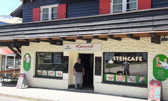 Ein traditionelles Restaurant mit einem einladenden Eingang und einer Terrasse. Die Fassade ist in hellen und dunklen Farben gestaltet, mit einer kleinen Gruppe von Menüs und Angeboten auf Plakaten. | © Alpengrill Riezlern | Veronika Senn