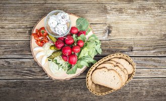 A rustic wooden board with fresh vegetables, radishes, and pickled cucumbers. Beside it, there is a basket with slices of whole grain bread. | © Alpenhotel DAS KÜREN