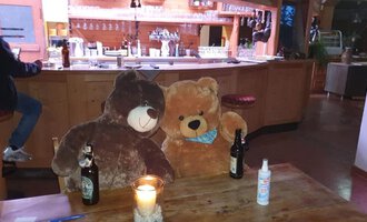 Two large teddy bears are sitting at a table in a bar. On the table are two beer bottles and a candle.