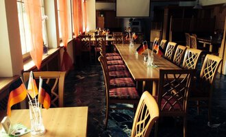 An inviting restaurant with long wooden tables and chairs. Small German flags are placed on the tables, and windows with red curtains create a cozy atmosphere.