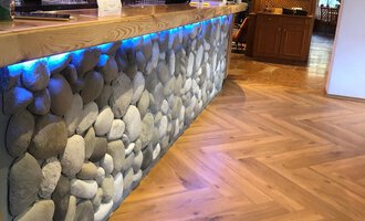 A stylish bar with a stone element and a lit wooden surface. The floor features an attractive herringbone pattern.