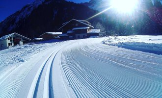 A snowy landscape with groomed ski tracks and a sunny sky. In the background, cabins and mountains can be seen.