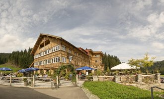 Ein rustikales Hotel mit Holzfassade in einer schönen Naturlandschaft. Der Außenbereich hat Sitzgelegenheiten und Sonnenschirme unter einem bewölkten Himmel. | © Auenhütte | Yorck Dertinger