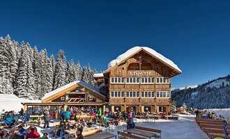 Ein gemütliches Bergrestaurant umgeben von schneebedeckten Bäumen. Viele Menschen genießen die Winteratmosphäre draußen auf der Terrasse. | © Auenhütte | Yorck Dertinger