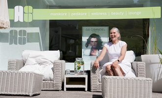 Eine entspannte Frau sitzt in einem modernen Lounge-Bereich mit geflochtenen Möbeln. Im Hintergrund sind Elemente für Wellness und Schönheit sichtbar. | © Annett Schwärzler Nails & Spa | Stefanie Haid