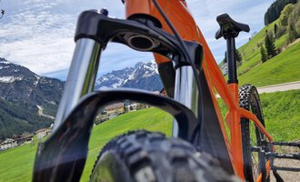© Austrian Ski- & Service Ranch | Trixi Leitner Ein orangefarbenes Mountainbike steht auf einem grünen Hügel mit Blick auf die Berge. Der Himmel ist klar und es ist ein sonniger Tag. | © Austrian Ski- & Service Ranch | Trixi Leitner