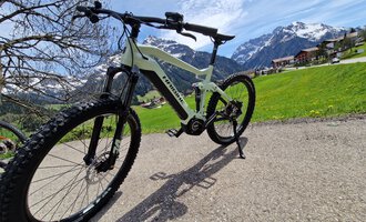 © Austrian Ski- & Service Ranch | Trixi Leitner Ein modernes Mountainbike steht auf einem Weg inmitten einer malerischen Berglandschaft. Im Hintergrund sind schneebedeckte Berge und grüne Wiesen zu sehen. | © Austrian Ski- & Service Ranch | Trixi Leitner