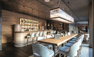 An elegant interior of a modern restaurant with a long wooden table and stylish chairs. In the background, there is a well-lit bar with a variety of drinks. | © Travel Charme Ifen Hotel