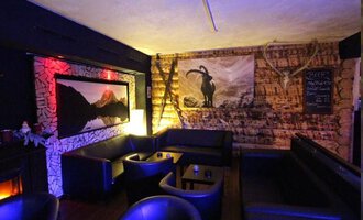 A cozy interior with dark leather furniture and wooden paneling. On the wall hangs a picture of a mountain panorama and a stylized goat motif. | © Heiko Möller m&m bar