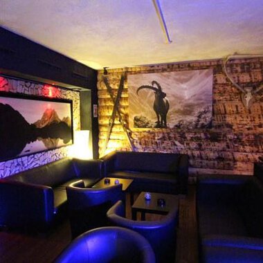 Ein gemütlicher Innenraum mit dunklen Ledermöbeln und einer Holzverkleidung. An der Wand hängt ein Bild eines Bergpanoramas und ein stilisiertes Ziegenmotiv. | © Heiko Möller m&m bar