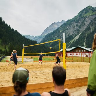 Ein Beachvolleyballspiel in den Bergen, umgeben von einer malerischen Landschaft. Die Spieler sind aktiv auf dem Sandplatz, während Zuschauer zuschauen. | © Kleinwalsertal Tourismus | Oliver Farys