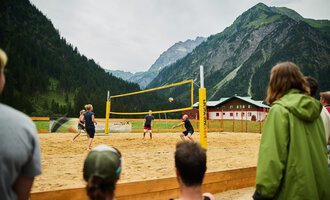 Ein Beachvolleyballspiel in den Bergen, umgeben von einer malerischen Landschaft. Die Spieler sind aktiv auf dem Sandplatz, während Zuschauer zuschauen. | © Kleinwalsertal Tourismus | Oliver Farys