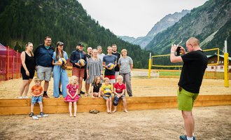 Eine Gruppe von Menschen steht auf einem Sandplatz mit Volleyballnetz in den Bergen. Jemand macht Fotos von ihnen, während sie lächeln und posieren. | © Kleinwalsertal Tourismus | Oliver Farys