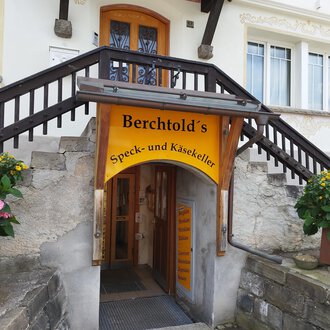 An entrance area to Berchtold's bacon and cheese cellar. Above the door hangs a yellow sign, surrounded by flowers. | © Kleinwalsertal Tourismus | N. Lughammer
