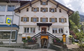A traditional alpine building with many windows and a curved entrance. Colorful flowers adorn the stairs in front of the house. | © Kleinwalsertal Tourismus | N. Lughammer