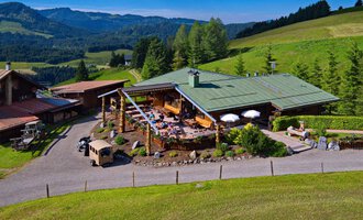 Eine malerische Almhütte umgeben von grünen Wiesen und Wald. Auf der Terrasse genießen Menschen die sonnige Aussicht in die Berge. | © Bergstüble | Jürgen Waffenschmidt