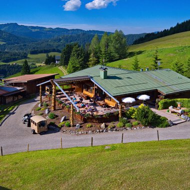 A picturesque mountain hut surrounded by green meadows and woods. On the terrace, people enjoy the sunny view of the mountains. | © Bergstüble | Jürgen Waffenschmidt