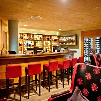 © Hotel Birkenhöhe | Fotograf Krug Eine gemütliche Bar mit einem langen Holztheken und roten Barstühlen. Im Hintergrund sind Regale mit Getränken und eine moderne Uhr sichtbar. | © Hotel Birkenhöhe | Fotograf Krug