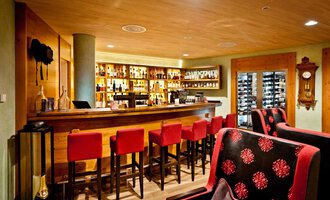 © Hotel Birkenhöhe | Fotograf Krug Eine gemütliche Bar mit einem langen Holztheken und roten Barstühlen. Im Hintergrund sind Regale mit Getränken und eine moderne Uhr sichtbar. | © Hotel Birkenhöhe | Fotograf Krug