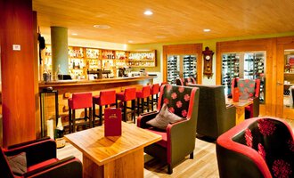 © Hotel Birkenhöhe | Fotograf Krug Eine gemütliche Bar mit einem Holzinterieur und roten Stühlen. Die Beleuchtung ist warm und einladend, mit Regalen voller Getränke im Hintergrund. | © Hotel Birkenhöhe | Fotograf Krug