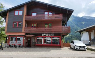 Ein modernes Gebäude mit einem Restaurant namens "sweetchili" im Erdgeschoss. Im Hintergrund sind grüne Berge und ein blauer Himmel zu sehen. | © Kleinwalsertal Tourismus | N. Lughammer