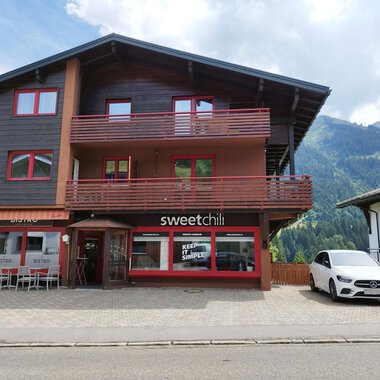 Ein modernes Gebäude mit einem Restaurant namens "sweetchili" im Erdgeschoss. Im Hintergrund sind grüne Berge und ein blauer Himmel zu sehen. | © Kleinwalsertal Tourismus | N. Lughammer