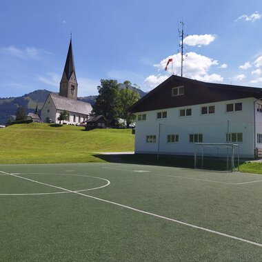Ein Fußballplatz vor einem modernen Gebäude mit einer Kirche im Hintergrund. Die Umgebung ist grün mit Hügeln und einem klaren blauen Himmel. | © Kleinwalsertal Tourismus | N. Lughammer