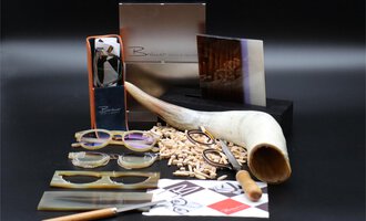 Ein stilvolles Arrangement aus Brillen, Holzstücken und Werkzeugen. Im Hintergrund sind elegante Verpackungen und eine große Hornschale zu sehen. | © Bräuer Optik & Akustik | AlpenMedia-Design