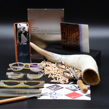 Ein stilvolles Arrangement aus Brillen, Holzstücken und Werkzeugen. Im Hintergrund sind elegante Verpackungen und eine große Hornschale zu sehen. | © Bräuer Optik & Akustik | AlpenMedia-Design