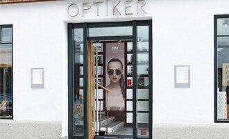 Ein Optikerladen mit großen Fenstern und einer auffälligen Werbefigur. Die Eingangstür ist geöffnet und lädt Kunden ein. | © Bräuer Optik & Akustik | AlpenMedia-Design