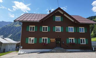© Kleinwalsertal Tourismus | Nicole Lughammer Ein traditionelles altes Haus mit brauner Fassade und grünen Fensterläden. Im Hintergrund sind Berge und ein blauer Himmel sichtbar. | © Kleinwalsertal Tourismus | Nicole Lughammer