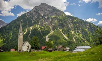 © Kleinwalsertal Tourismus | Steffen Berschin Eine malerische Landschaft mit einem Dorf und einer beeindruckenden Bergkulisse. Im Vordergrund steht eine alte Kirche und saftige Wiesen. | © Kleinwalsertal Tourismus | Steffen Berschin