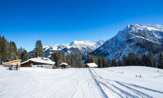 Eine schneebedeckte Landschaft mit Bergen und klar blauem Himmel. Im Vordergrund sind einige Hütten und Skifahrer zu sehen. | © Bühlalpe | Frank Drechsel
