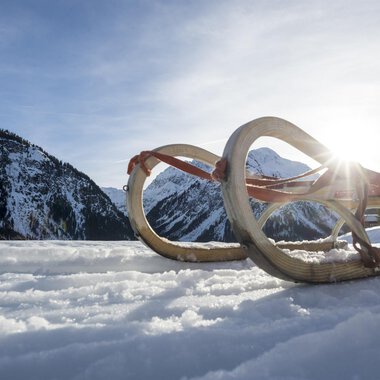 Ein Schlitten liegt im Schnee, umgeben von einer winterlichen Berglandschaft. Die Sonne strahlt hinter den Bergen hervor. | © Kleinwalsertal Tourismus | Dominik Berchtold
