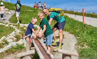 Vater und seine zwei Söhne spielen auf einem Weg im Freien. Im Hintergrund sind weitere Personen zu sehen, und die Umgebung ist grün und sonnig. | © OBERSTDORF · KLEINWALSERTAL BERGBAHNEN