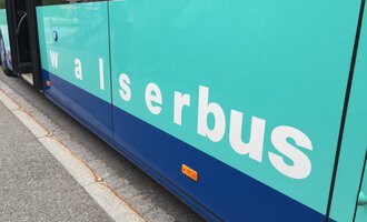 A bus with the inscription "walserbus" in blue and turquoise colors. The bus is standing at a stop on a paved road. | © Kleinwalsertal Tourismus