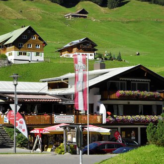 A charming inn with blooming balconies in a green mountain landscape. In the background, more traditional cottages can be seen. | © Cafe Baad Grund | Roman Schuster